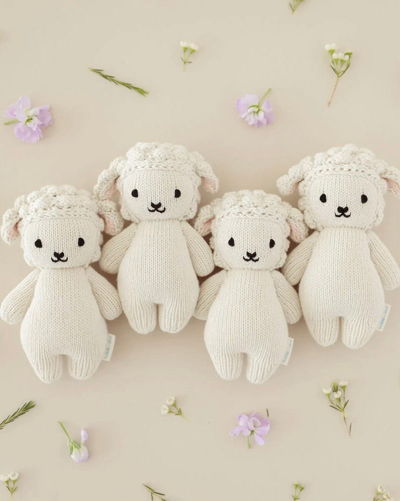 Four Cuddle + Kind Baby Lambs, hand-knit from Peruvian cotton yarn, with white bodies and cream faces, each adorned with pink floral crowns, arranged symmetrically on a beige background scattered with small.