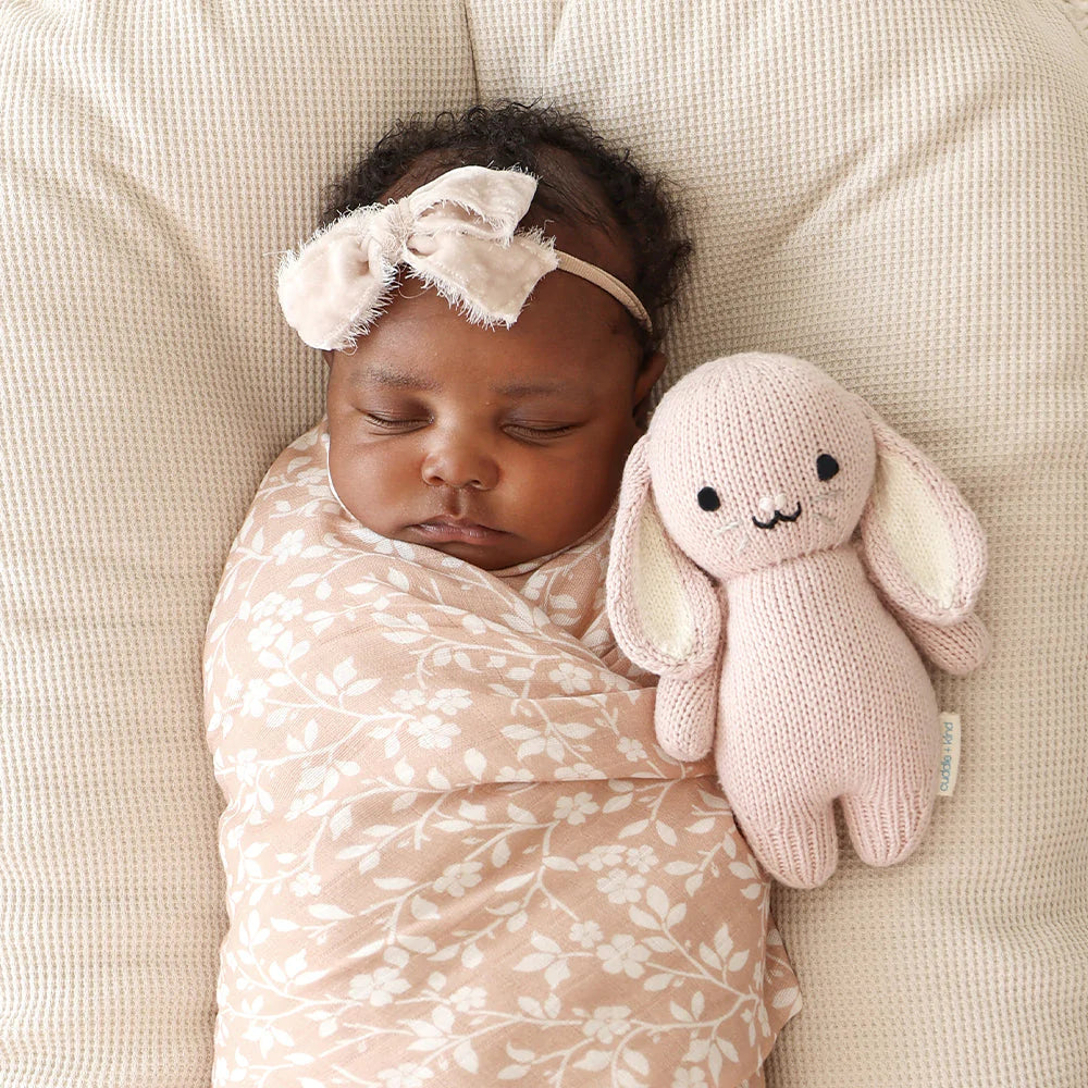 A newborn baby swaddled in a floral blanket sleeps peacefully next to a Cuddle + Kind Baby Bunny, with a delicate bow headband on her head.