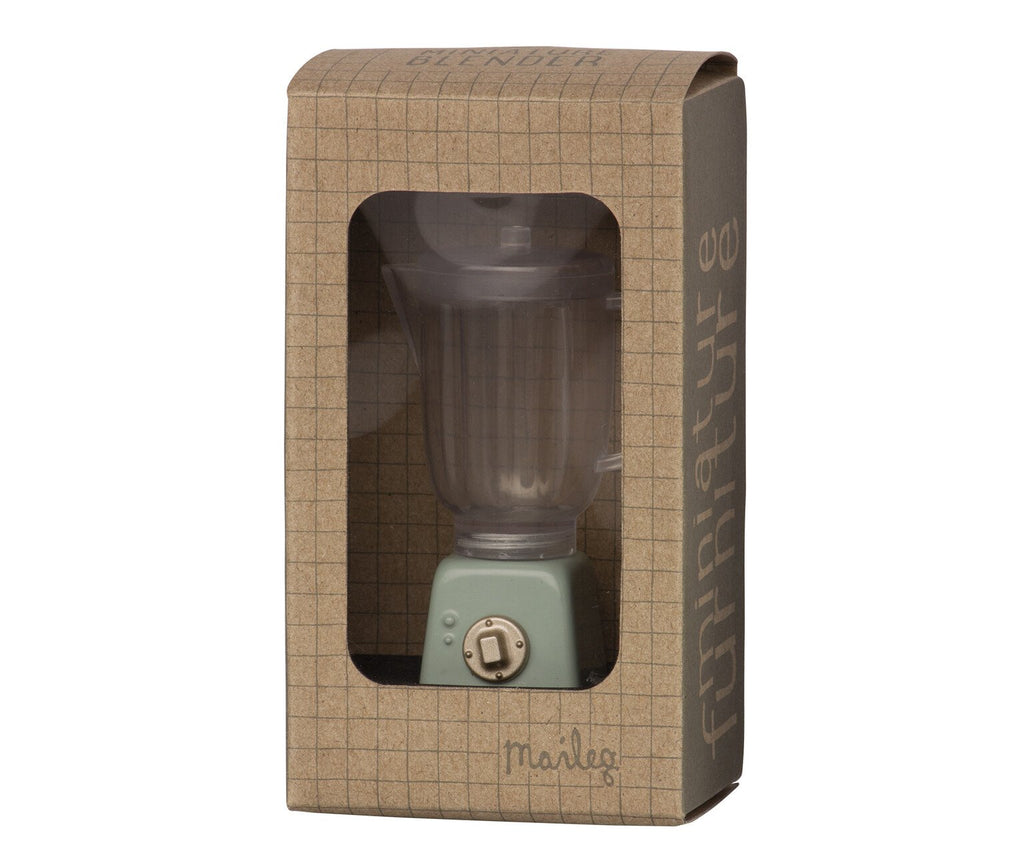 A small, mint green Maileg Miniature Blender is displayed in brown grid-patterned cardboard packaging with a clear plastic window. Perfect for children's toy collections and pretend play, the blender features a dial on the front. The packaging has text that reads "Maileg" and another text design along the side.