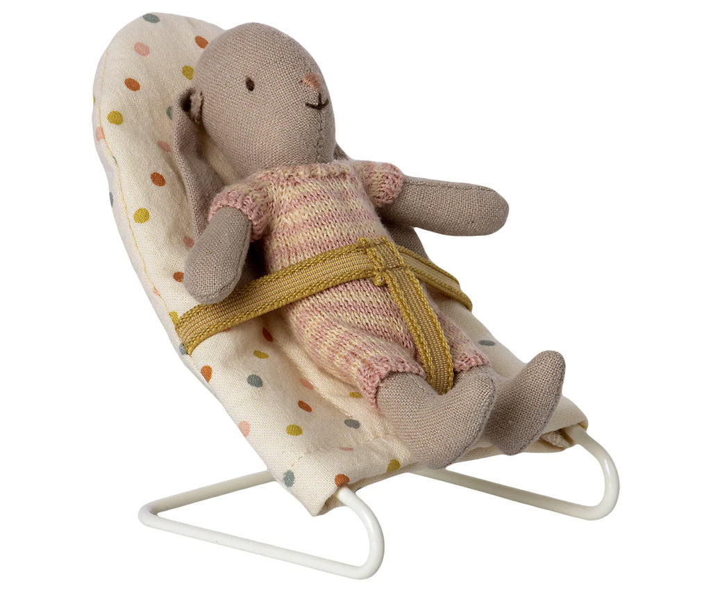 A small stuffed bear wearing a knitted outfit is sitting in a Maileg Micro Babysitter with multicolored polka dots. The babysitter is secured with a yellow belt and has a white metal frame. The bear's arms and legs are spread out comfortably, looking as cozy as sleepy wakey mice snuggled in their little beds.