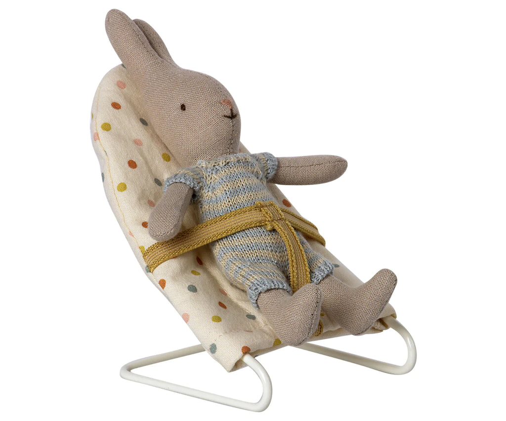 A My sized Maileg rabbit toy wearing a blue and yellow knitted outfit is seated in a Maileg Micro Babysitter. The chair has a safety belt around the bunny and rests on a simple white metal frame.