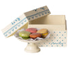 A hand-painted cake stand holds five colorful Maileg Macarons displayed in front of an open cream-colored box with a blue floral and dotted design. The box lid is propped open behind the platter, showcasing the French pastry-themed treat.