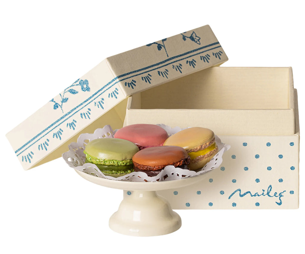 A hand-painted cake stand holds five colorful Maileg Macarons displayed in front of an open cream-colored box with a blue floral and dotted design. The box lid is propped open behind the platter, showcasing the French pastry-themed treat.