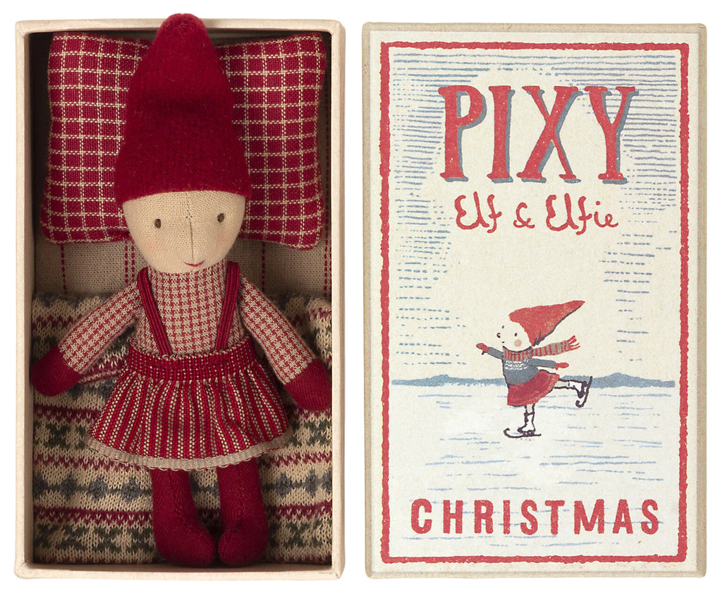 A small doll dressed in a red hat, red and white sweater, and a red skirt lies in a box with a red and white checked pillow and a striped blanket. The box reads "Maileg Pixy Elfie," complete with an image of an elf ice skating—a perfect Christmas friend for your Maileg ginger bread house.