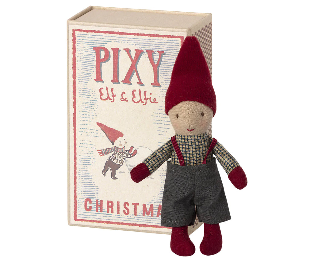 A small doll dressed as an elf stands beside a box labeled "Maileg Pixy Elfie." The elf wears a red hat, plaid shirt, dark overalls, and red mittens and socks. The box features an illustration of a similar elf and festive text alongside a Maileg ginger bread house.