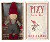 A small figurine of an elf with a red hat, red gloves, and gray overalls is in a box. The box cover reads "Maileg Pixy Elfie," featuring an elf rolling a snowball. This charming Christmas friend is lying on a patterned pillow and blanket, perfect for your Maileg ginger bread house.