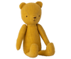 A mustard yellow stuffed Maileg Teddy Junior with simple black stitching for facial features sits upright against a plain white background. The bear, part of the Maileg Teddy Junior family, has a minimalist design with jointed arms and legs, giving it a charming vintage look.