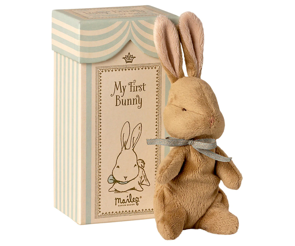 A soft plush bunny with a bow around its neck sits next to a decorative rectangular box labeled "Maileg My First Bunny - Light Blue," perfect as a baby shower gift. The box showcases a charming drawing of a bunny and features a striped pastel design.