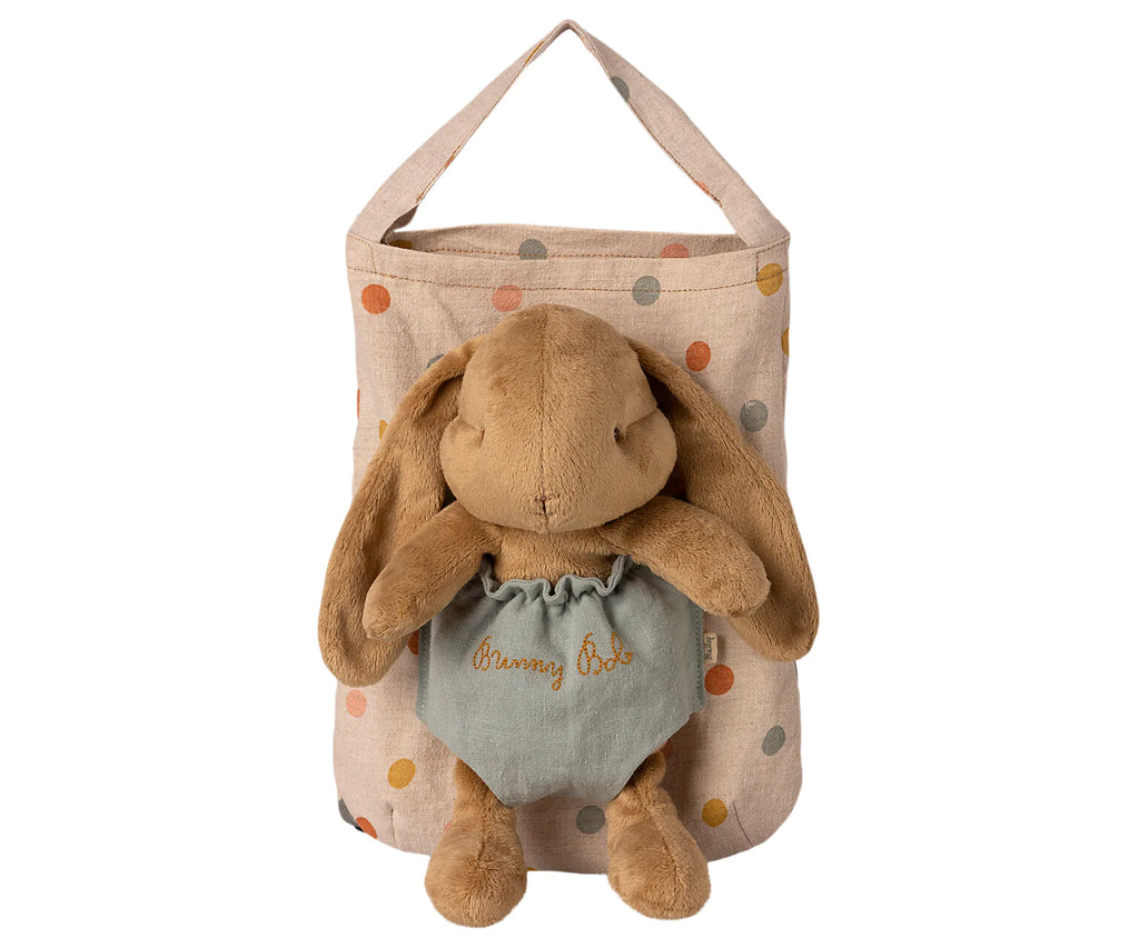 A Maileg Bunny Bob with droopy ears, dressed in a green outfit embroidered with "Bunny Bob," is placed inside a beige fabric polka dot bag. The pouch, adorned with multi-colored polka dots and equipped with a handle for hanging, adds an extra touch of charm.