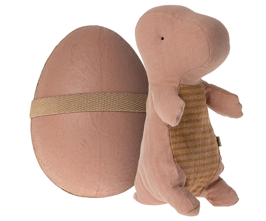 The Maileg Small Gantosaurus in Egg - Rose stands next to its matching egg-shaped carrying case. The dinosaur, which is light pink with a brown striped belly, has soft and cuddly fabric. The egg case is secured with a tan strap, and both items have a gentle plush appearance, making them perfect for lifelike dino enthusiasts.