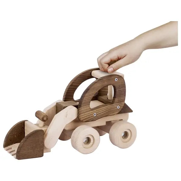 A child's hand playing with a Goki Bulldozer on a white background. The toy, made of sustainable raw material, consists of a cab and detachable trailer, both crafted from natural and dark stained wood.