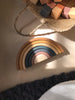 A Handmade Rainbow Stacker - Mustard, a non-toxic wooden toy with pastel-colored arcs, is placed under natural sunlight on a wooden surface. Beside it, there's a woven basket containing various wooden blocks. A knitted dark gray blanket partially appears in the bottom of the image.