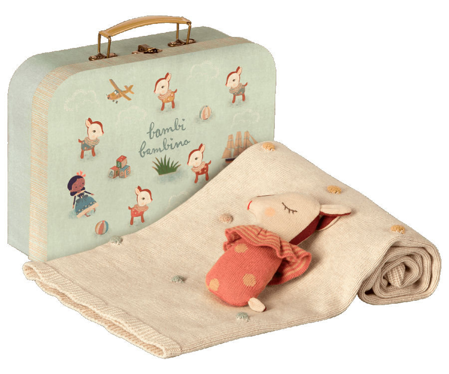 A pale green suitcase with whimsical illustrations of children playing is paired with a beige knitted blanket adorned with small pom-poms. A Bambi rattle, resembling a sleepy animal wearing an orange outfit, lies partially wrapped in the blanket. This Maileg Baby Gift Set is perfect for any little one.