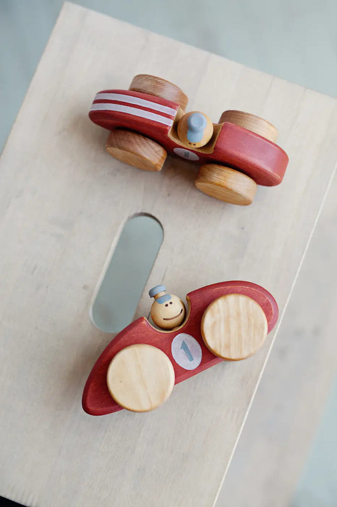 Two Handmade Wooden Toy Race Cars sit on a light-colored wooden surface. The toy car on the left has red stripes, while the one on the right is red with a white number "1" on the side. Made from sustainably harvested birch wood, both cars have round wooden wheels and small wooden drivers with hats.