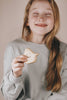 A girl with long, light brown hair is smiling with her eyes closed. She is wearing a light gray sweatshirt and holding a small Wooden Ghost Emotions toy crafted from natural linseed oil. The scene captures pure basic emotions of joy and serenity.