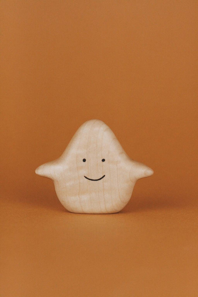 A small, cute Wooden Ghost Emotions figure with a simple, smiling face and outstretched arms is set against a solid orange background. The light-colored figure, treated with natural linseed oil, showcases a smooth texture that evokes basic emotions of joy and warmth.