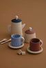 A Raduga Grez Handmade Wooden Tea Set consisting of a teapot, a sugar bowl, two mugs, two saucers, two small spoons, and three small wooden blocks. The items are crafted from light wood with the mugs in blue and brown. The set is arranged on a plain, tan surface.