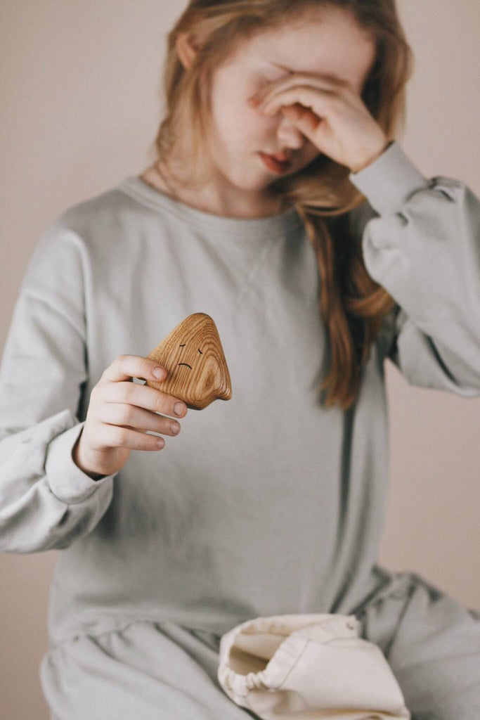 A person wearing a light grey top is sitting and holding a Wooden Ghost Emotions figure with an unhappy face carved on it. The figure appears to be treated with natural linseed oil. The person is wiping or rubbing their face with their other hand, while the background remains neutral and unobtrusive.