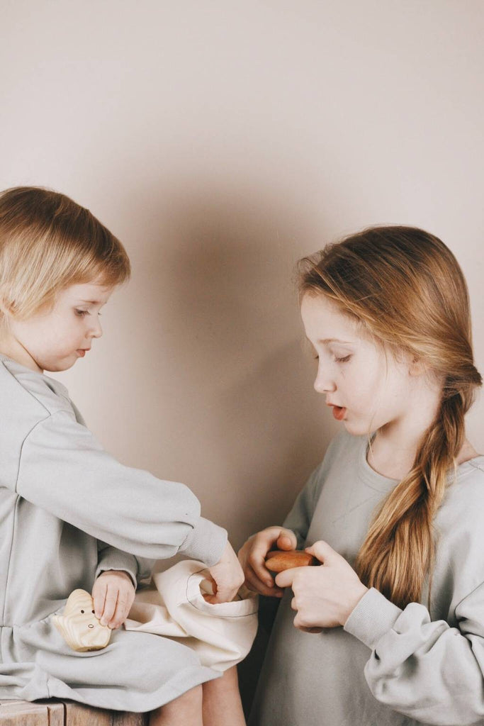 Two children are engaged in an activity. The younger child on the left, with short blonde hair, is reaching into a small cloth bag. The older child on the right, with long blonde hair in a braid, is holding a Wooden Ghost Emotions figure and looking down. Both children are wearing greyish-blue outfits.