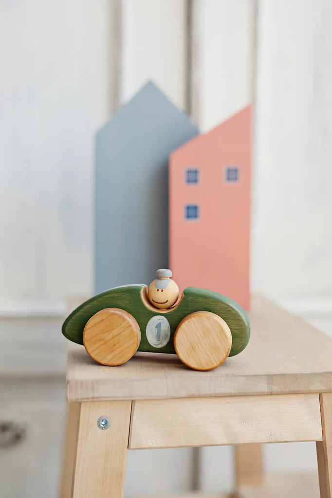 A Handmade Wooden Toy Race Car made from sustainably harvested birch wood boasts a green body and light brown wheels. It features a painted number "1" and a small figurine driver with a hat. In the background, two wooden house shapes are painted blue and pink.