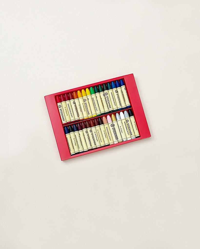 A box of vibrant Stockmar Wax Stick Crayons neatly arranged in rows on a plain background, with labels facing upwards for easy identification of colors.