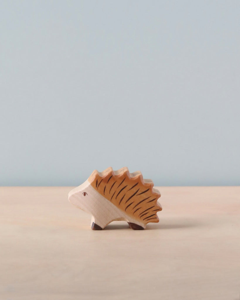 A small, wooden Holztiger Hedgehog, reminiscent of HOLZTIGER figures, is placed on a light wooden surface with a plain light blue background. The handcrafted wood toy is simplistic in design with carved details representing its spines and facial features. Made in Europe, it captures timeless charm.