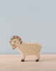 A Holztiger Ram, this wooden toy ram with intricately carved details stands on a light-colored surface against a pale blue background. The ram has curved horns and a textured body, encapsulating the whimsical and handmade appeal typical of handcrafted wood toys made in Europe.