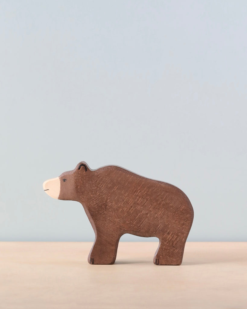 A Holztiger Brown Bear with a simplistic, minimalist design stands on a light wooden surface against a plain light blue background. Reminiscent of HOLZTIGER figures, the handcrafted wood bear is painted brown with lighter brown detailing on its face and paws.