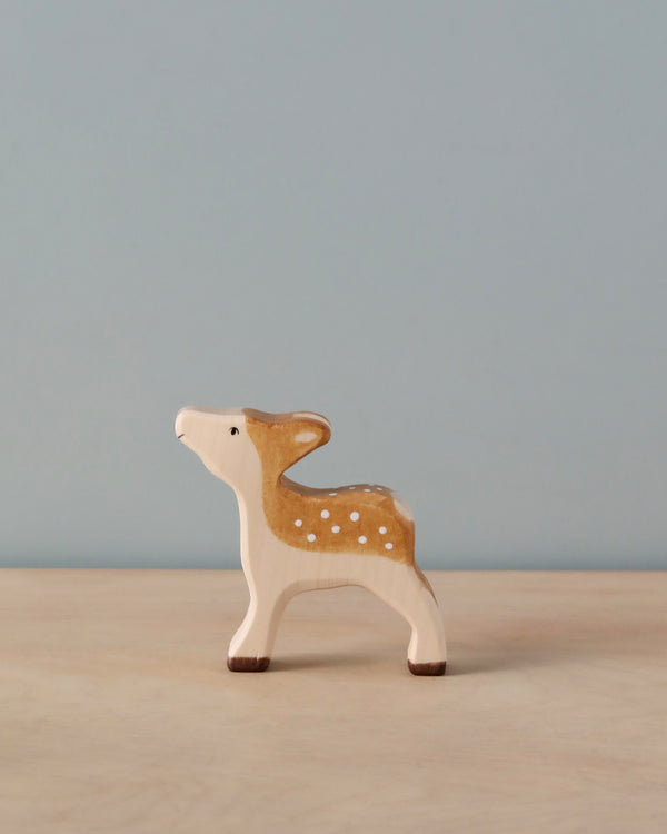 A small, handcrafted wooden Holztiger Fawn stands on a light wooden surface. Made in Europe by HOLZTIGER figures, the fawn is painted with light brown and white colors, featuring white spots on its back. The background is plain and light blue.
