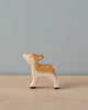 A small, handcrafted wooden Holztiger Fawn stands on a light wooden surface. Made in Europe by HOLZTIGER figures, the fawn is painted with light brown and white colors, featuring white spots on its back. The background is plain and light blue.
