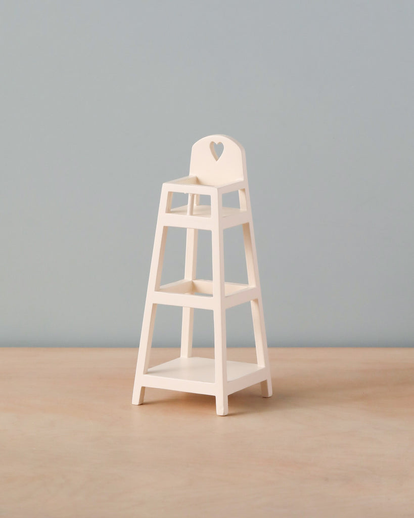 A small, white wooden Maileg My High Chair for children is placed on a light-colored wooden surface. This charming piece of nursery furniture features a heart-shaped cutout on the backrest and boasts a simple, minimalist design against a plain background.