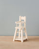 A small, white, wooden Maileg Micro High Chair sits empty on a light wood surface against a plain, light blue background. Perfect for micro-sized Maileg friends or tiny bunnies and rabbits, the high chair features a tray, a footrest, and a simple, classic design.