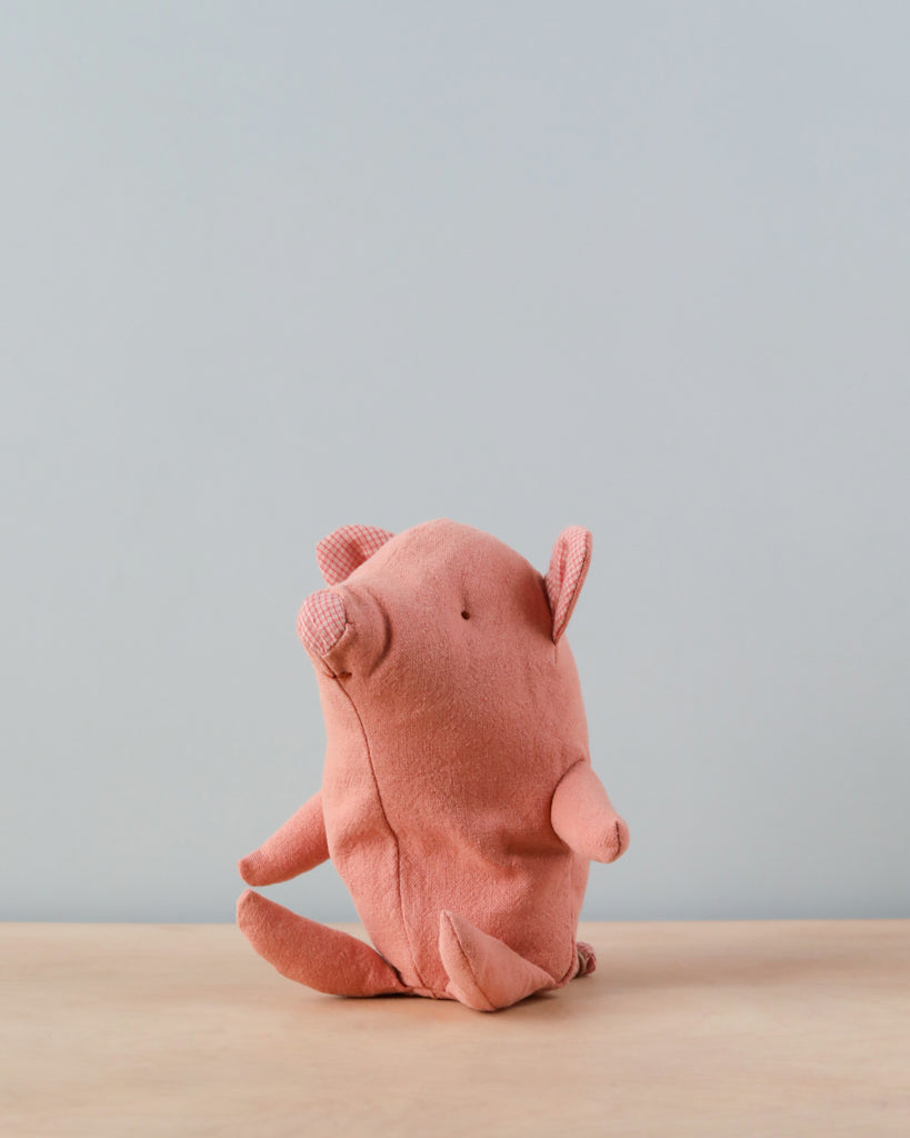 Maileg Truffles the Pig, Baby, a small plush toy made from light pink recycled polyester fabric, sits on a wooden surface against a plain gray background. Shaped like an adorable pig with simple features including a snout, ears, and tiny limbs, it boasts a minimalist yet charming appearance.