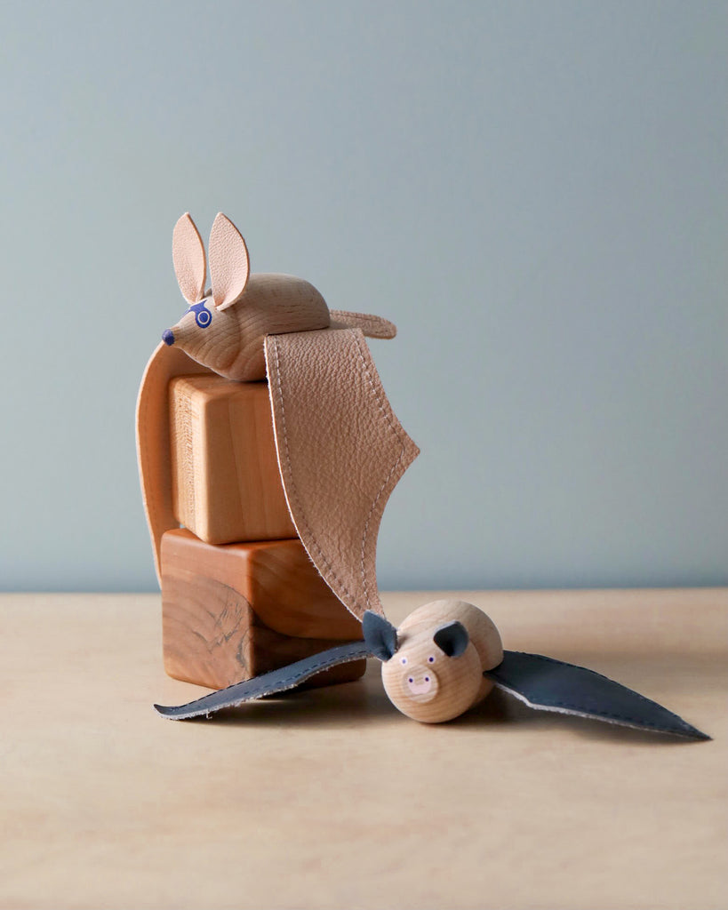 A Handmade Wooden Bat sculpture with leather wings and a smaller bat lying flat, both with simplified features and visible wood grain, on a neutral background.