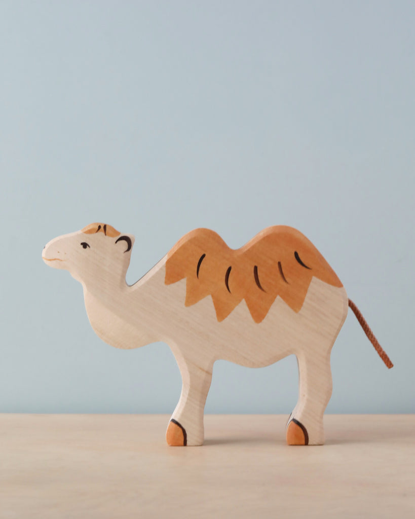 A handcrafted **Holztiger Camel** with a minimalist design stands on a light-colored surface against a pale blue background. Made in Europe, the camel features two humps painted in light brown, matching its legs and hooves. This charming piece is reminiscent of HOLZTIGER figures.