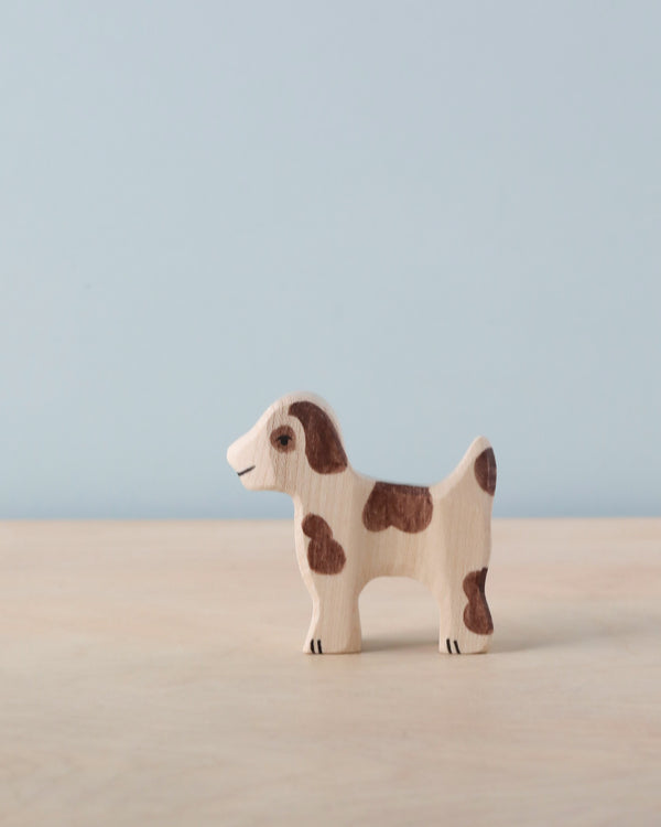 A small, handcrafted wood Holztiger Small Farm Dog with a simple design. The dog is painted light brown with dark brown spots and stands on a smooth, light-colored wooden surface against a plain blue background. Made in Europe, this charming piece mirrors the quality of HOLZTIGER figures.