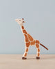 A small, wooden Holztiger Small Feeding Giraffe with a light brown body and dark brown spots stands on a neutral-colored surface against a light blue background. Handcrafted wood and part of the HOLZTIGER figures collection, this giraffe has a simple, minimalist design and is positioned in profile, facing left.