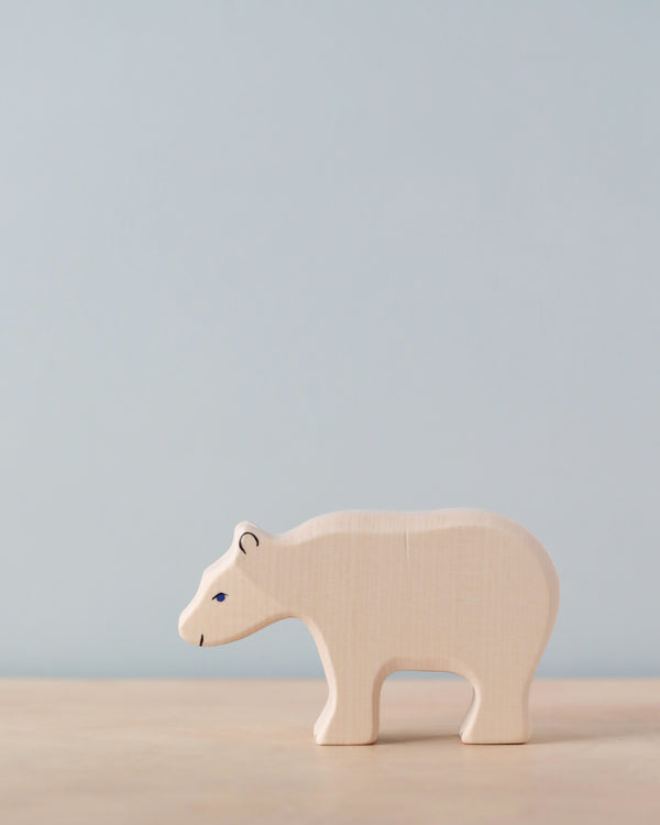 A small, light-colored wooden bear figurine stands on a light wooden surface against a plain, light blue background. The handcrafted Holztiger Polar Bear, Feeding has minimalistic features, with simple painted eyes and nose. As part of the celebrated HOLZTIGER figures collection, it proudly boasts its Made in Europe origin.