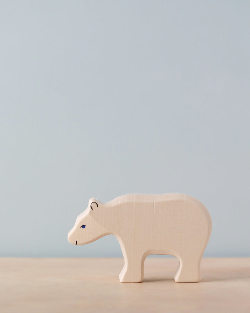 A small, light-colored wooden bear figurine stands on a light wooden surface against a plain, light blue background. The handcrafted Holztiger Polar Bear, Feeding has minimalistic features, with simple painted eyes and nose. As part of the celebrated HOLZTIGER figures collection, it proudly boasts its Made in Europe origin.