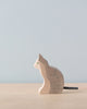 A small wooden cat figurine with a light natural wood finish and subtle painted details sits on a beige surface against a plain light blue background. The high-quality, handcrafted wood design of this Holztiger Sitting Gray Cat figure showcases the cat facing left, with its tail slightly curled behind it.