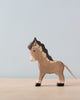 A small handcrafted wood toy donkey stands on a light wooden surface against a plain light blue background. The high-quality Holztiger Small Donkey figure is painted brown with black accents for details such as the mane, hooves, and tail. It has a simple, charming design.