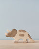 A Holztiger Triceratops stands on a light wooden surface against a pale blue background. The painted toy, made in Europe, is primarily beige with gray spots, featuring three horns and the characteristic frill of a triceratops.