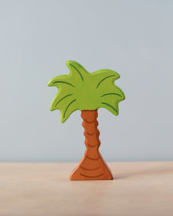 A small, wooden, hand-painted palm tree figurine with a brown trunk and green fronds stands upright on a light-colored surface against a light blue background. This delightful piece is part of the handcrafted Holztiger Palm Tree collection, proudly made in Europe.