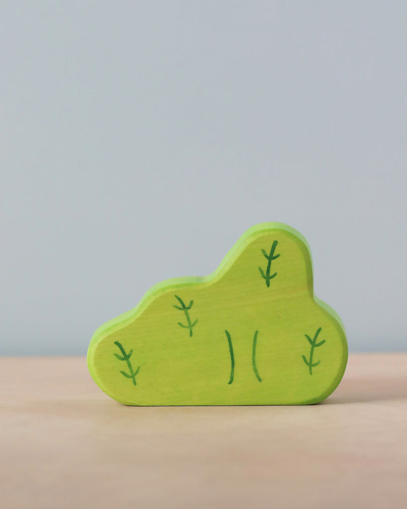 A small, green wooden toy shaped like a bush or tree stands on a light brown surface against a gray background. Handcrafted in Europe and part of the charming Holztiger Bush collection, the toy is painted with simple dark green lines to represent leaves and branches.