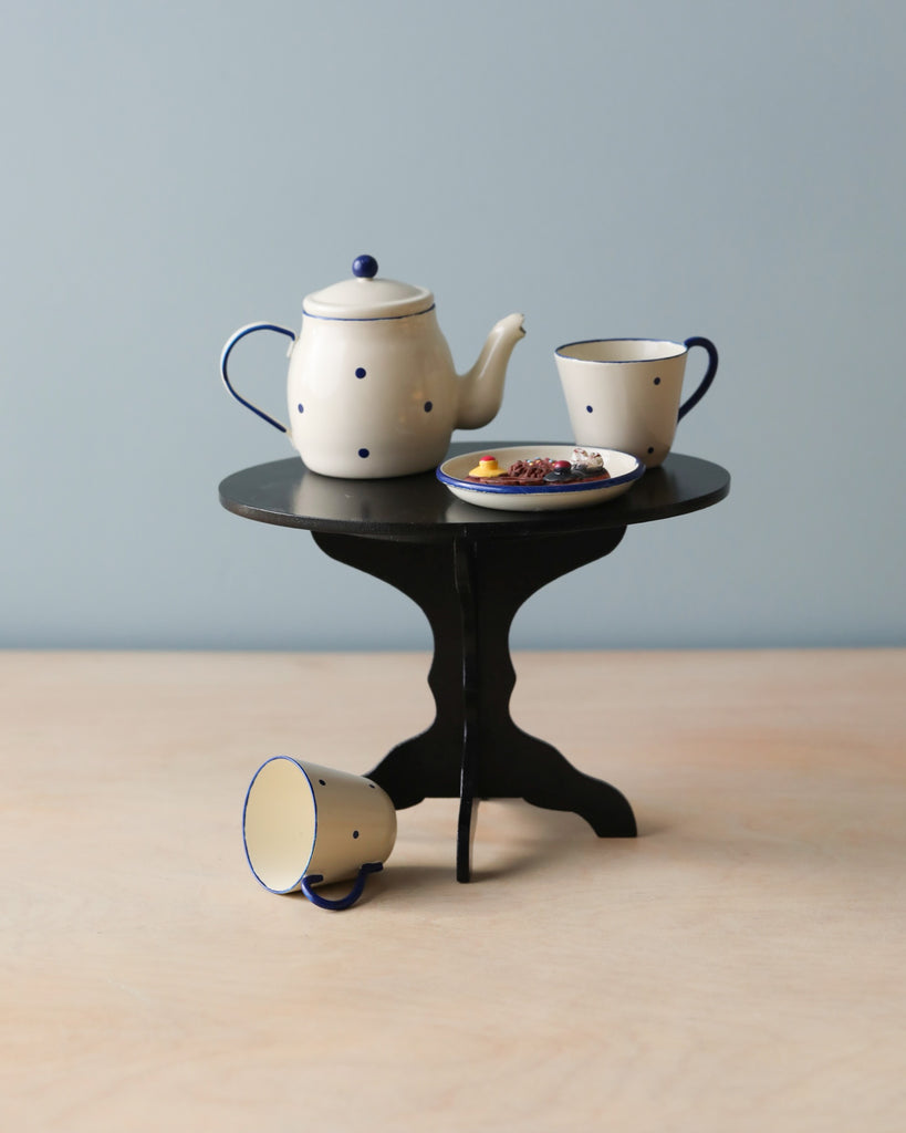 A Maileg | Miniature Tea & Biscuits for Two set with a white teapot featuring blue dots and two matching cups, arranged on a small round black table, with one cup tipped over on the floor beside it.