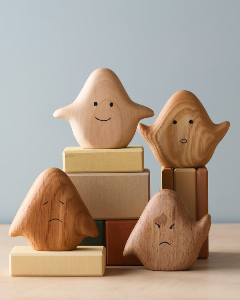 Image of Wooden Ghost Emotions, four small wooden ghost figures with different facial expressions, treated with natural linseed oil. They sit on and around a stack of pastel-colored wooden blocks. The expressions include happy, sad, angry, and surprised, depicting basic emotions.