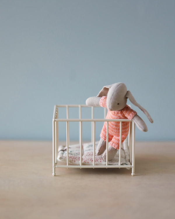 A small My-sized Maileg stuffed bunny wearing an orange knitted outfit stands propped up inside a white miniature Maileg Playpen with a cute cushion and a blanket lying on the playpen's floor. The background is a soft blue wall, and the scene is set on a light-colored wooden surface.