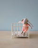 A small My-sized Maileg stuffed bunny wearing an orange knitted outfit stands propped up inside a white miniature Maileg Playpen with a cute cushion and a blanket lying on the playpen's floor. The background is a soft blue wall, and the scene is set on a light-colored wooden surface.