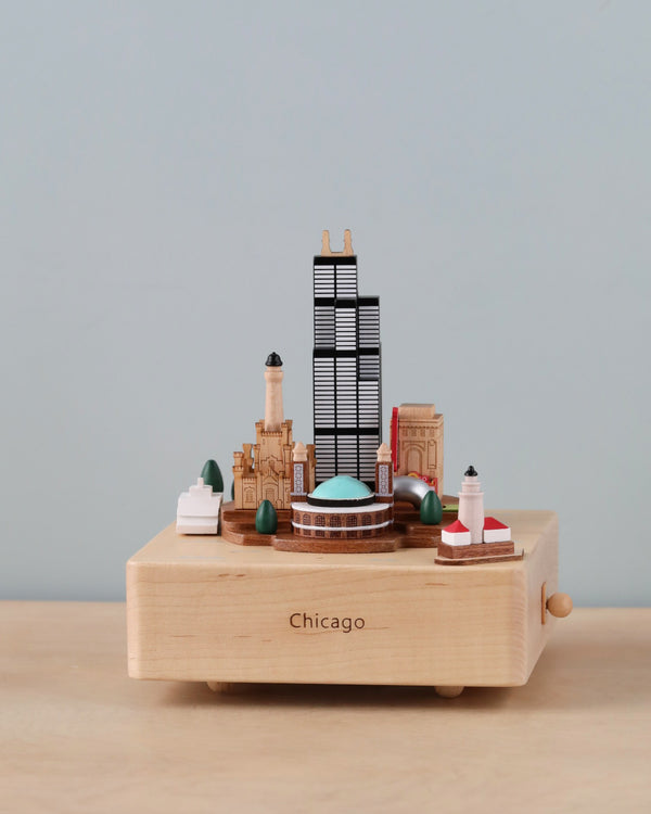 A miniature Chicago Wooden Music Box model of Chicago landmarks on a platform labeled "Chicago," featuring iconic buildings and a lighthouse, set against a neutral background.