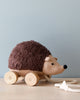 Hedgehog Pull Toy, a plush toy with wooden wheels and a pull string, rests on a light wooden surface against a light blue background. Made with organic cotton, Hedgehog Pull Toy features a brown textured back and a beige face, with a white string ending in a star for pulling—a perfect companion from the Enchanted Forest.